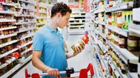 A man stands between shelves in a supermarket. He is holding a jar of olives in one hand and looking at it. His other hand is on the handle of the shopping cart.