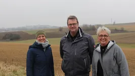 Three people in front of an empty field. A hill with several buildings can be seen in the background.